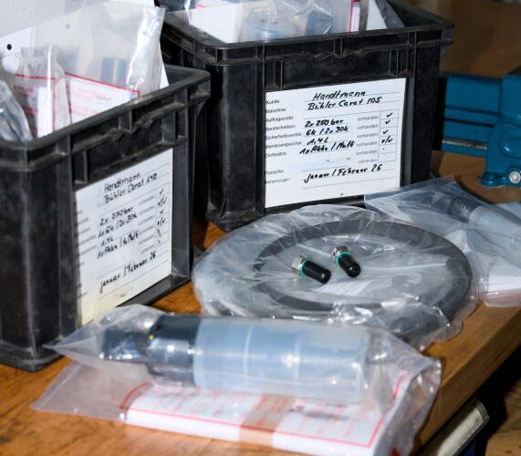 Zwei schwarze Kunststoffkisten mit Ersatzteilen und Dokumentation, daneben verpackte Ersatzteile wie ein Ring